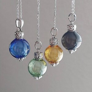 May include: Four glass pendant necklaces with silver-colored chains and decorative caps. The pendants are round and come in blue, green, yellow, and black. The necklaces are hanging against a neutral background.