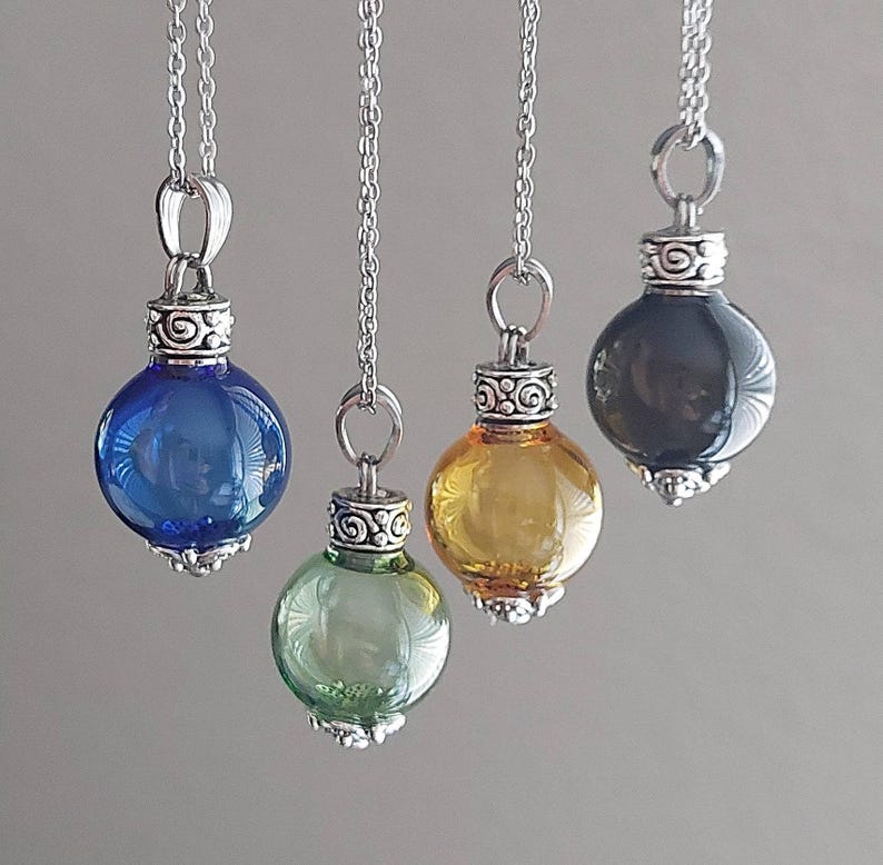 May include: Four glass pendant necklaces with silver-colored chains and decorative caps. The pendants are round and come in blue, green, yellow, and black. The necklaces are hanging against a neutral background.