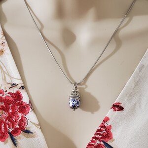 Puede incluir: Un collar de cadena de plata con un colgante de porcelana azul y blanco. El colgante tiene forma de bellota y tiene un borde plateado.