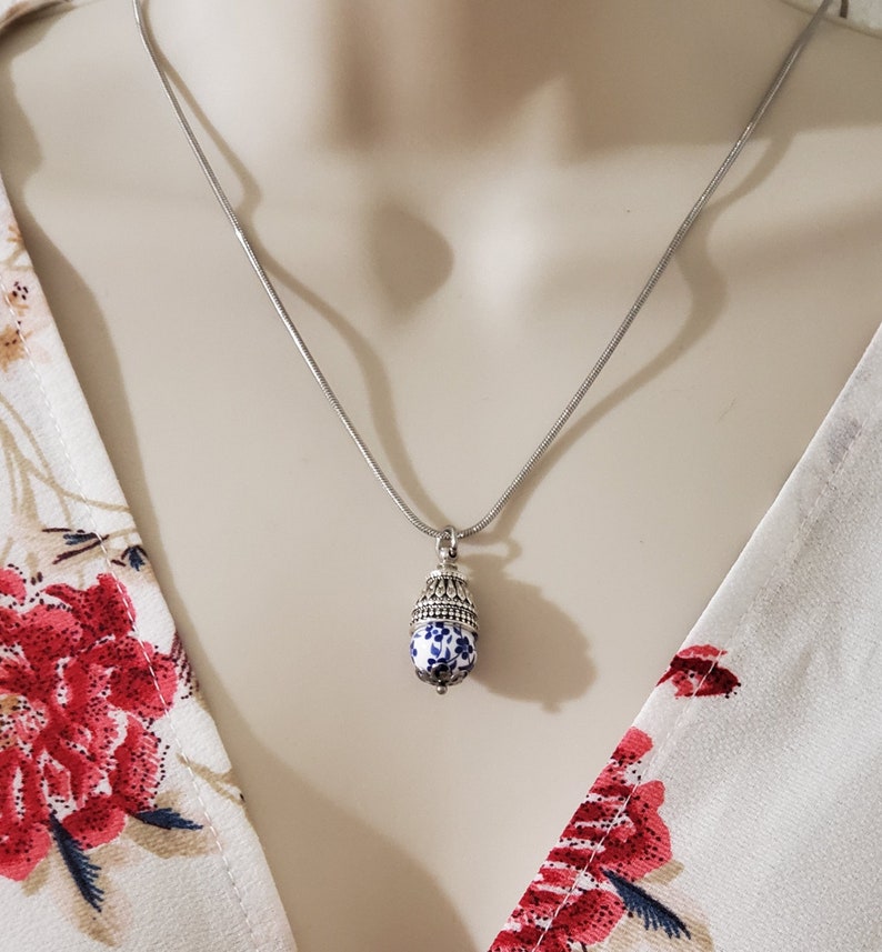 Puede incluir: Un collar de cadena de plata con un colgante de porcelana azul y blanco. El colgante tiene forma de bellota y tiene un borde plateado.