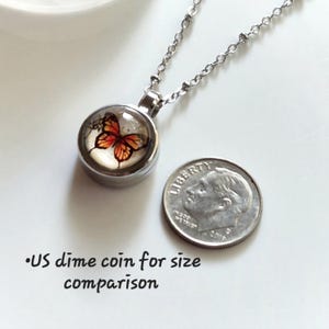 Puede incluir: Collar con colgante plateado con un dije redondo con un dise&ntilde;o de mariposa en tonos naranja y negro. El collar se muestra junto a una moneda de diez centavos estadounidense para comparar el tama&ntilde;o.