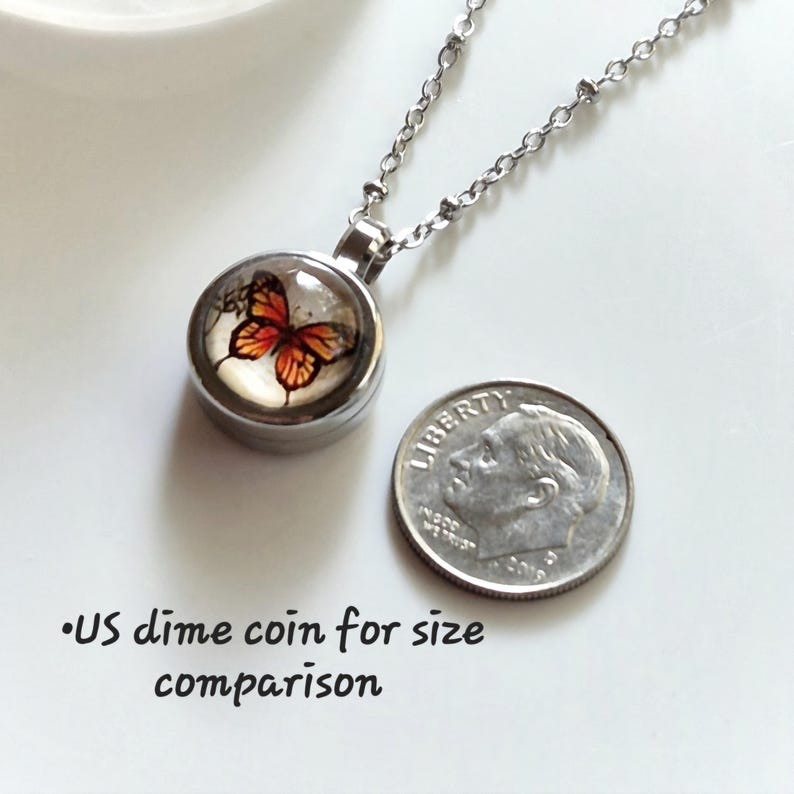 Puede incluir: Collar con colgante plateado con un dije redondo con un dise&ntilde;o de mariposa en tonos naranja y negro. El collar se muestra junto a una moneda de diez centavos estadounidense para comparar el tama&ntilde;o.