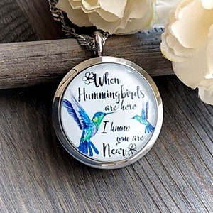 May include: Silver-toned pendant necklace with a round charm featuring a hummingbird design. The charm has the text "When Hummingbirds are here I know you are Near" and illustrations of hummingbirds in shades of blue and green.