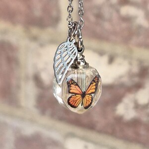 May include: A silver-toned necklace featuring a clear, spherical pendant containing a monarch butterfly graphic. A small, silver-toned wing charm hangs from the same jump ring as the pendant, suspended by a delicate silver-toned metal chain.