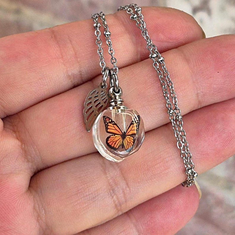 May include: A silver-toned necklace featuring a clear, spherical glass pendant containing a miniature orange and black monarch butterfly graphic. A small, silver-toned metal wing-shaped charm hangs beside the glass sphere on a delicate chain.