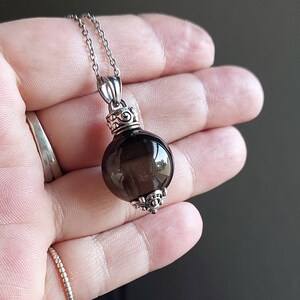 May include: A silver necklace featuring a dark, translucent, round pendant. The pendant is attached to a delicate silver chain. The pendant has decorative silver accents at the top and bottom. The necklace is displayed against a neutral background.