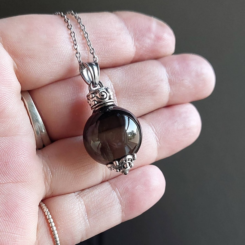 May include: A silver necklace featuring a dark, translucent, round pendant. The pendant is attached to a delicate silver chain. The pendant has decorative silver accents at the top and bottom. The necklace is displayed against a neutral background.