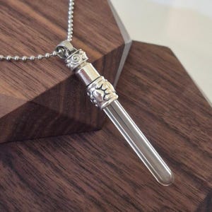 May include: A silver-colored pendant necklace featuring a clear glass vial. The vial is encased in a decorative silver metal setting with ornate detailing. The necklace chain is a delicate ball chain.