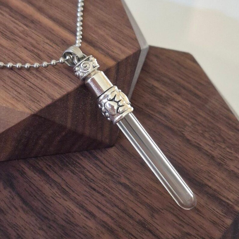 May include: A silver-colored pendant necklace featuring a clear glass vial. The vial is encased in a decorative silver metal setting with ornate detailing. The necklace chain is a delicate ball chain.
