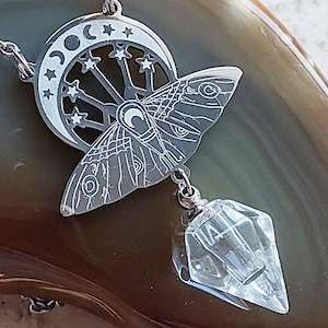 May include: A silver-toned metal pendant necklace featuring a moth silhouette with intricate wing patterns, a crescent moon, and star cutouts. A clear, faceted, diamond-shaped crystal drop hangs from the bottom of the moth-shaped metal piece.