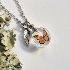 May include: A silver-toned necklace featuring a clear, hexagonal pendant containing an orange and black monarch butterfly graphic, accompanied by a small silver-toned wing charm suspended from a delicate metal chain link necklace.