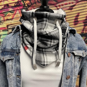 Puede incluir: Un calentador de cuello a cuadros en blanco y negro con cordones blancos, exhibido en un maniquí. El cuello está superpuesto a una camisa blanca y una chaqueta vaquera con botones color cobre. El fondo presenta arte de graffiti colorido.