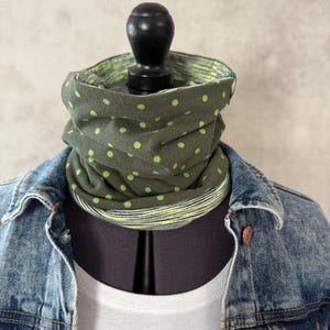 Könnte beinhalten: Olivgrüner Rundschal mit hellgrünen Punkten. Der Schal ist wendbar und hat innen ein Streifenmuster. Der Schal wird auf einer Schaufensterpuppe mit Jeansjacke und weißem T-Shirt präsentiert.