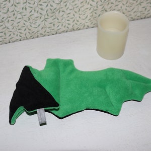 Pode incluir: Um morcego de pelúcia verde e preto com textura de feltro. O morcego está deitado sobre uma superfície branca.