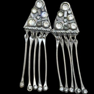 May include: A pair of silver-toned triangular earrings featuring a textured metal surface embedded with multiple clear round rhinestones. Each earring has four long, slender dangling metal strands, with each strand tipped by a small clear rhinestone.