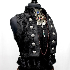 May include: A black velvet vest with silver buttons and black tassels. The vest has a high collar and is worn over a black top with a red beaded necklace and silver jewelry.