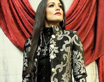 May include: A black and silver brocade jacket with a high collar and a black skirt. The jacket has a floral pattern and is worn over a black shirt with a silver cross pendant.