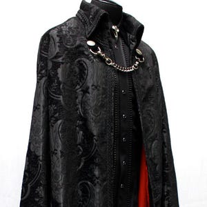 May include: A black cloak with a paisley pattern and a red lining. The cloak has a high collar and is fastened with a silver chain. The garment is displayed on a black mannequin.