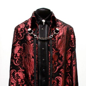 May include: A long, red and black brocade cape with a black trim and a silver chain necklace. The cape is on a black stand.