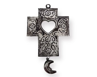 Puede incluir: Una cruz de metal plateado con un recorte en forma de corazón y diseños florales. Un amuleto de media luna cuelga de la parte inferior de la cruz.