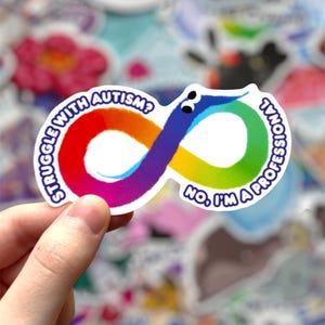 Pode incluir: Um autocolante colorido com um símbolo de infinito arco-íris e o texto "Struggle with Autism?" e "No, I'm a Professional". O autocolante tem uma borda branca e é segurado por uma mão, com outros autocolantes no fundo.