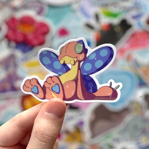 May include: A sticker of a cartoon creature with a pink and yellow body, blue wings with white polka dots, and blue heart-shaped feet. The creature has large green eyes and a small mouth.