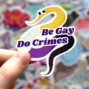 May include: A sticker featuring a stylized worm with a yellow head, white body, purple midsection, and black tail. The sticker has the text "Be Gay Do Crimes" in purple. The background is filled with other colorful stickers.