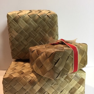 May include: Three woven boxes made of natural materials, stacked on top of each other. The top box has a red ribbon tied around it.