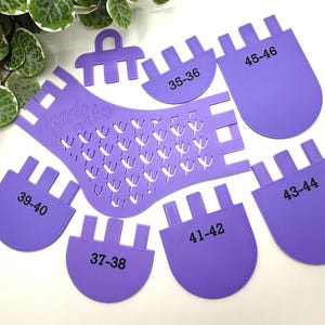 May include: A collection of purple shoe size measuring tools. The set includes a foot-shaped template with heart cutouts, and several size markers labelled with numbers. The tools are arranged on a white surface with green foliage in the background.