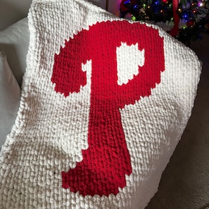 May include: A white, chunky-knit blanket featuring a large red letter "P" in the center. The blanket is folded over a gray pillow, with a Christmas tree visible in the background. The blanket is made of a soft, textured material.