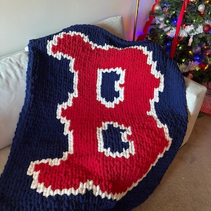 May include: A navy blue chunky knit blanket with a large red letter "B" and white trim. The blanket is draped over a white sofa. This cosy throw is a decorative home accessory.