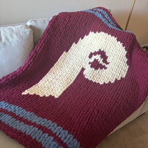 May include: A burgundy and cream-colored, chunky knit blanket with a large cream-colored letter "P" design. The blanket has two light blue stripes near the bottom. The blanket is draped over a light-colored couch.