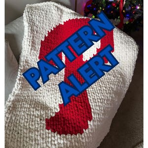 Puede incluir: Una manta de punto grueso en crema con un gran dise&ntilde;o rojo. La manta tiene un aspecto texturizado y est&aacute; doblada. Las palabras "PATTERN ALERT" se muestran en texto azul en la manta.