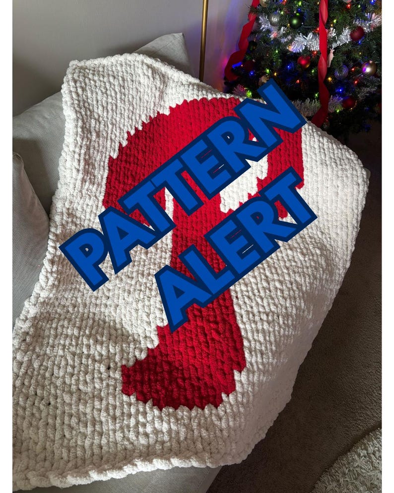 Puede incluir: Una manta de punto grueso en crema con un gran dise&ntilde;o rojo. La manta tiene un aspecto texturizado y est&aacute; doblada. Las palabras "PATTERN ALERT" se muestran en texto azul en la manta.