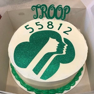 May include: A round white cake with green frosting and a green glitter logo featuring two silhouettes. The cake is decorated with the word "TROOP" and the number "55812" in green. The cake is in a white box.