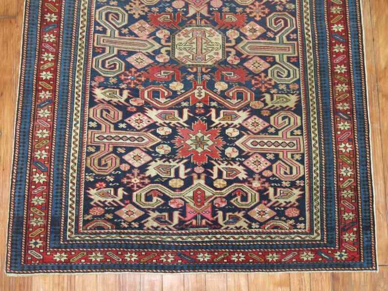 K&ouml;nnte beinhalten: Ein traditioneller Teppich mit dunkelblauem Hintergrund und komplizierten geometrischen Mustern in Rot-, Rosa-, Beige- und Gr&uuml;nt&ouml;nen. Der Teppich hat einen Rand mit einem sich wiederholenden Muster aus Sternen und Blumen.