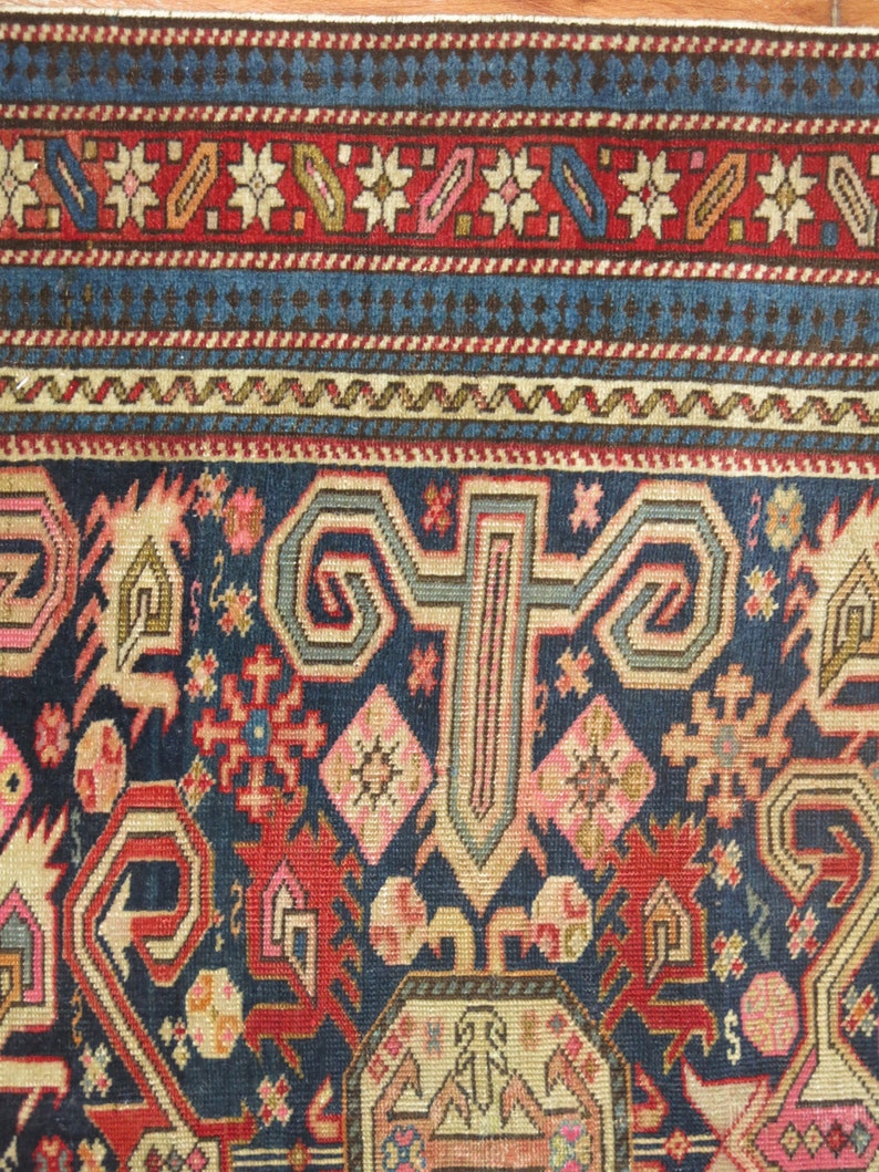 K&ouml;nnte beinhalten: Eine Nahaufnahme eines traditionellen Teppichs mit komplizierten Mustern in Rot-, Blau- und Cremet&ouml;nen. Der Teppich hat ein zentrales Medaillon mit einem geometrischen Muster und floralen Motiven.