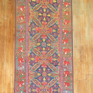 May include: A long, narrow rug with a traditional Persian design. The rug has a dark blue background with intricate patterns in shades of red, yellow, green, and white. The rug is bordered by a red and green floral pattern.