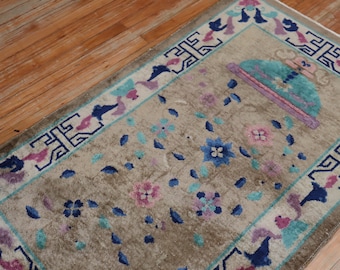 Op de afbeelding: Een beige kleed met een blauw, roze en groen bloemenpatroon. Het kleed heeft een rand met een geometrisch ontwerp in blauw en wit.