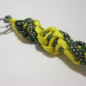 May include: A handmade keychain featuring a braided design. The keychain is made of yellow and black-green paracord, with a silver-colored split ring for attaching keys. The braided section is approximately 4 inches long.