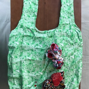 May include: A green reusable shopping bag with a white floral pattern. The bag is folded and has two fabric handles. There are two small fabric pouches inside the bag, one with a red rose design and the other with a multi-colored floral design.