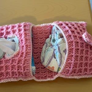 May include: A pink crocheted diaper caddy with a textured waffle stitch design. The caddy has a compartment for diapers and a separate compartment with a wipe dispenser. The caddy is designed to hold baby essentials.
