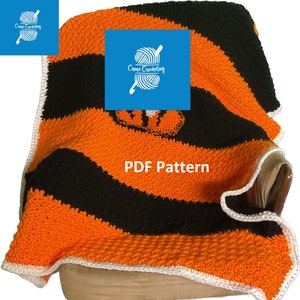 Könnte beinhalten: Eine gehäkelte Decke mit orangefarbenen und schwarzen Streifen und einem weißen Rand. Die Decke ist gefaltet und über einen braunen Stuhl gelegt. Die Decke hat ein kleines orangefarbenes und schwarzes Tigerlogo in der Mitte. Der Text "PDF Pattern" ist auf die Decke gedruckt.