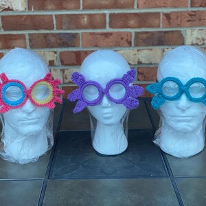 May include: Three pairs of handmade crochet glasses in vibrant colors. The glasses are displayed on white mannequin heads. One pair is pink, blue, and yellow. Another is purple, and the third is teal. The glasses have a whimsical, decorative design.
