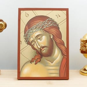 Può includere: Un'icona religiosa rettangolare raffigurante un ritratto dipinto di Gesù Cristo che indossa una corona di spine su uno sfondo dorato, incorniciata da un bordo rosso scuro, con il testo ATHOS ICONS HANDMADE GREEK ORTHODOX ICONS nell'angolo.