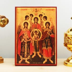 May include: A religious icon featuring figures with halos and robes, set against a gold background. The icon has a red border and includes text at the top and bottom. A wooden cross and a gold incense burner are in the foreground.