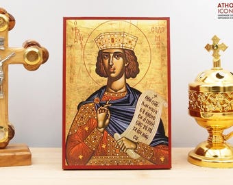May include: An Orthodox icon featuring a figure with a crown, holding a scroll with Greek text. The icon has a gold background and is framed in red. A wooden cross and a gold incense burner are also visible.