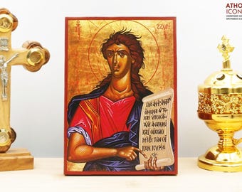 May include: A hand-painted icon of Saint John the Baptist holding a scroll with text in Greek. The icon is on a wooden background with a red border. The icon is a traditional Orthodox Christian icon.