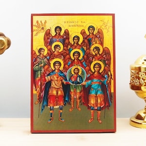 May include: A colorful religious icon depicting the Archangels. The icon is framed in a red wooden frame and features a gold background with a yellow halo around each angel. The text "Η ΣΥΝΑΞΙΣ ΤΩΝ ΑΡΧΑΓΓΕΛΩΝ" is written in Greek at the top of the icon.