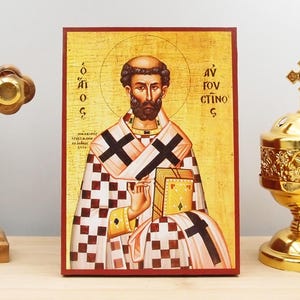 Può includere: Un'icona ortodossa raffigurante un santo con un'aureola, che tiene un libro. L'icona ha uno sfondo dorato e una cornice rossa. Il santo indossa una veste a scacchi con croci nere. Un incensiere d'oro e una croce di legno su entrambi i lati.